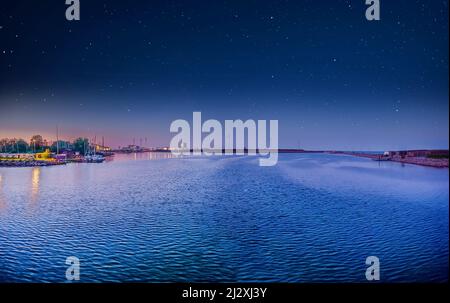 A bay with blue water, an grassland island Prøvestenen Syd with small houses, Wind turbines and a factory against the backdrop of a night sky with sta Stock Photo