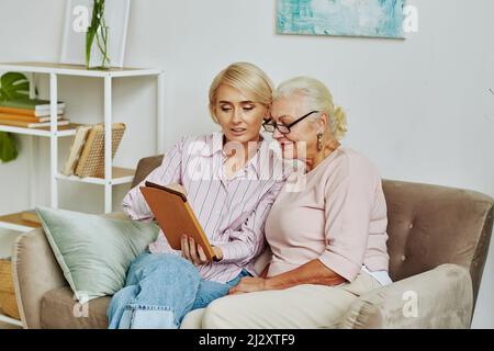 Portrait of blonde young woman helping senior mother using digital tablet and online services while sitting on couch at home together Stock Photo
