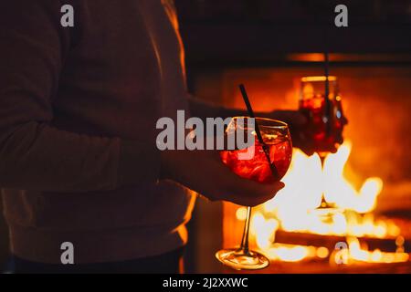 Crop unrecognizable male in casual shirt holding pair of glasses with alcohol Apperol Spritz cocktail with orange slices during romantic party by fire Stock Photo