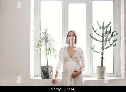 Young pregnant brunette woman touching her belly while standing near window wearing white dress Stock Photo