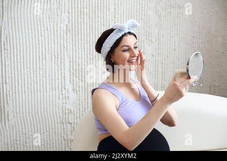 Happy beautiful pregnant brunette young woman applying cream in morning looking to the mirror surrounded by pants. Beauty and healthcare concept Stock Photo