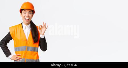 Satisfied happy asian female engineer, construction manager in safety helmet and reflective jacket showing okay gesture pleased, guarantee building Stock Photo