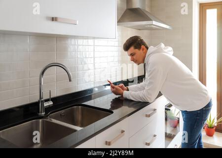 Young Caucasian man leaning over kitchen counter using mobile phone. Modern white kitchen with sink. White hoodie. European home interior. Stock Photo