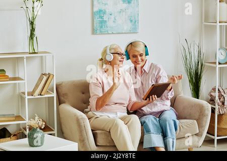 Portrait of smiling adult daughter with mother wearing headphones listening to music together and using digital tablet Stock Photo