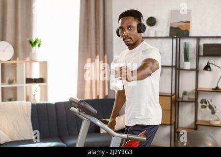 Way to health. Handsome african american man athlete preparing for cardio training on treadmill and listening to music with headphones looking at the Stock Photo