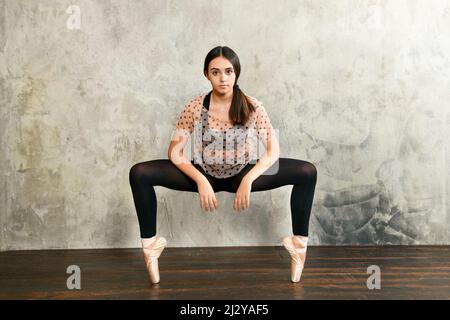 Front view of young ballerina posing en point balancing on the tips of her toes in special ballet shoes against a grunge grey studio wall looking quie Stock Photo