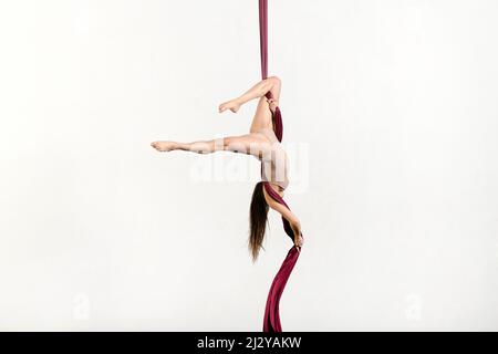 Full body side view of female aerialist in bodysuit dong falling stunt on aerial silks against white background in studio Stock Photo