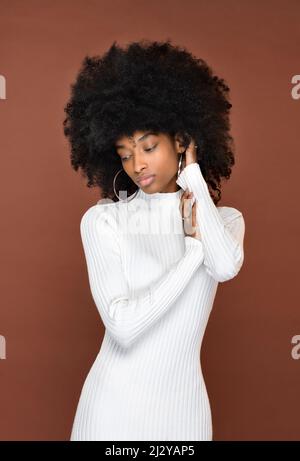 Slender calm Dominican lady with Afro hairstyle in white dress touching hair and looking down against brown background Stock Photo