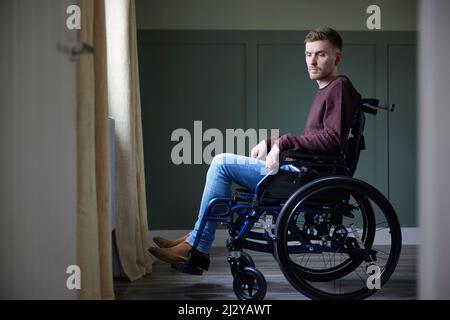 Depressed Young Man With Poor Mental Health In Wheelchair By Window At Home Stock Photo