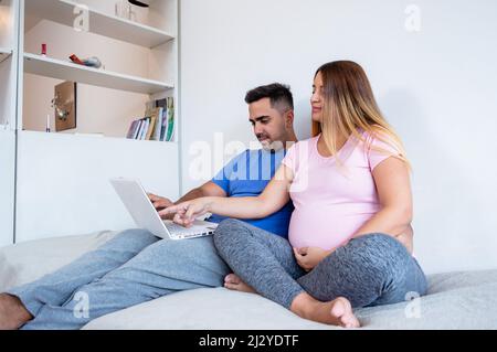 pregnant caucasian young woman in pink and gray pajamas, sitting on the bed with her husband, bearded man in blue pajamas, they are using the laptop, Stock Photo