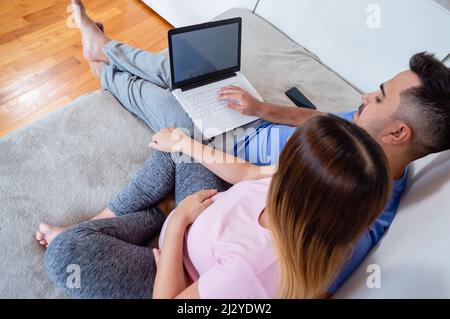 top rear view pregnant latin caucasian couple sitting on bed at home, watching laptop screen, using computer, surfing net, shopping online and watchin Stock Photo