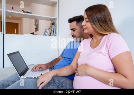 side view of pregnant caucasian young woman with her husband, bearded young man, in pajamas sitting on bed looking at laptop screen, technology concep Stock Photo