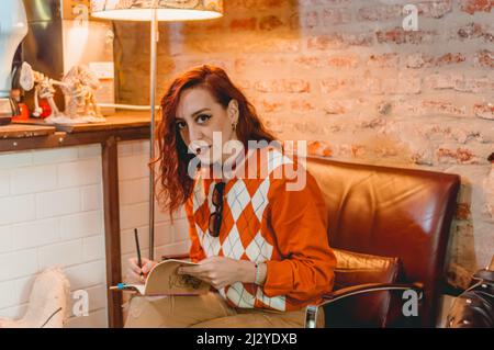 redhead caucasian young woman sitting on a sofa writing in a book in a restaurant surprised looking at the camera. Stock Photo