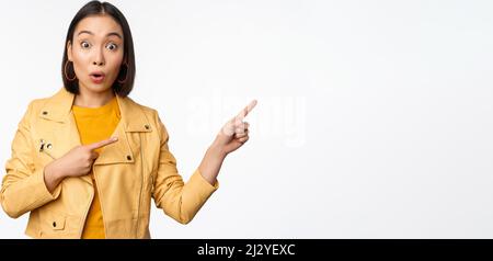 Enthusiastic asian girl pointing fingers right, showing advertisement and smiling, inviting to the store, standing over white background Stock Photo