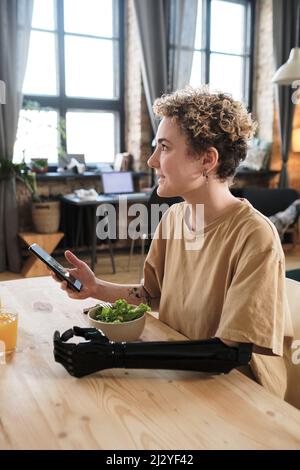 Young woman with prosthetic arm smiling at table while using her smartphone and having lunch at home Stock Photo