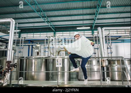Caucasian technician engineer man in uniform sitting and holding blueprint of industrial project with boiler and pipeline in beverage processing plant Stock Photo