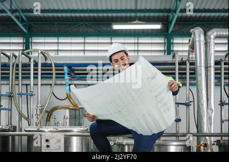 Caucasian technician engineer man in uniform sitting and holding blueprint of industrial project with boiler and pipeline in beverage processing plant Stock Photo