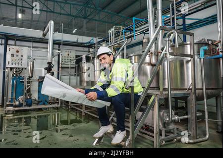 Caucasian technician engineer man in uniform sitting and holding blueprint of industrial project with boiler and pipeline in beverage processing plant Stock Photo