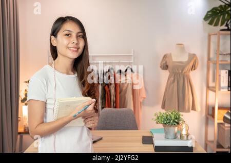 Beautiful asian online seller woman standing and holding notebook in clothing store at home office Stock Photo