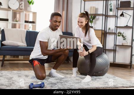 Workout at home. African American man and fitness trainer woman sitting after a workout on fitball having a video chat on laptop on the background of bright modern apartment. Stock Photo