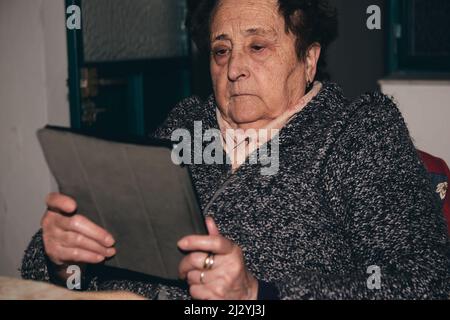 An elderly and surprised woman holding a tablet in her wrinkled hands. Stock Photo
