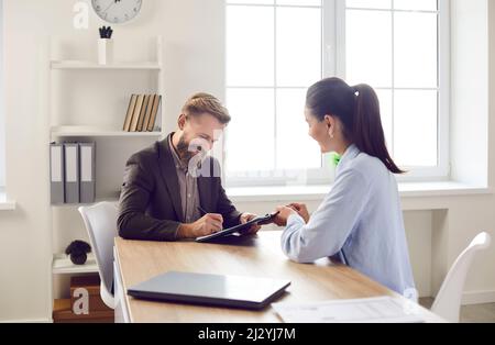 Happy man sitting at desk with bank worker and putting his signature on business document Stock Photo
