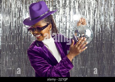 Cheerful young dark-skinned woman with shiny mirror disco ball in her hand on silver background. Stock Photo