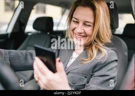 A young business woman drives a company car, checks her phone and smiling.. Stock Photo