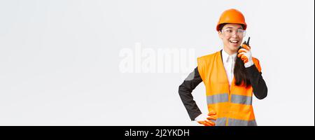 Confident pleased asian female engineer in safety helmet and uniform talking with chief architect using walkie-talkie. Satisfied construction Stock Photo