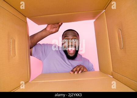 Low angel bottom view photo of funny excited guy wear purple t-shirt opening carton box isolated pink color background Stock Photo