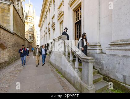 Town and Gown, contrast UK; a Cambridge University student emerges from the Senate House on her graduation day while local men walk past, Cambridge UK Stock Photo