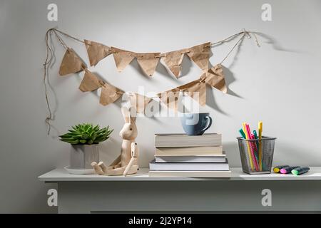 Echeveria in geometric pot, stack of books, pens in a pencil holder, felt-tip pens, wooden figurine of a hare are on shelf. Garland of craft paper fla Stock Photo