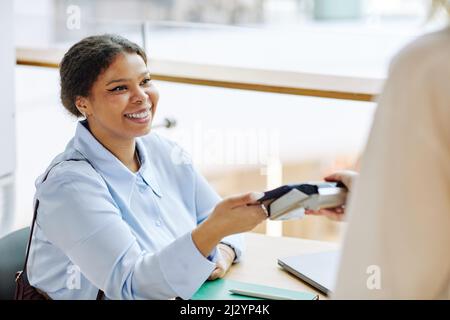 Portrait of smiling black woman paying with credit card in cafe and using bank payment machine, copy space