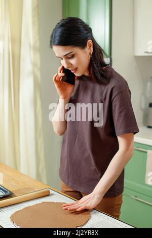 Portrait of young mestizo smiling woman talking on phone in home kitchen while cooking. Stock Photo
