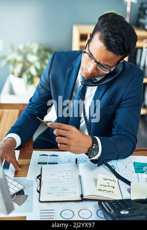 Good afternoon, how may I help. Shot of a focused young businessman seated at his desk while taking on the phone and typing on a keyboard in the Stock Photo