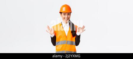 Architect made perfect plan. Confident asian female engineer in safety helmet showing okay gesture, have all under control, pleased with excellent Stock Photo