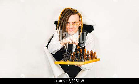 Young man in glasses with dreadlocks plays chess in hole of white background. Handsome male in white shirt, black vest and tie playing in logical boar Stock Photo
