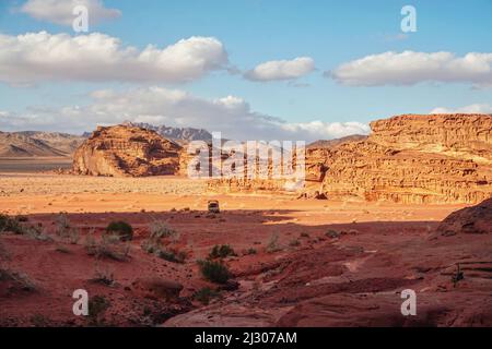 Rocky massifs on red sand desert, bright cloudy sky in background, small vehicle and camels at distance - typical scenery in Wadi Rum, Jordan Stock Photo