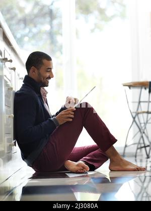 Theres always time for social media. Shot of a smiling young man sitting on his kitchen floor using a digital tablet. Stock Photo