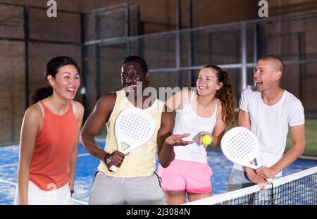 Laughing men and women with rackets and balls talking on indoor padel court Stock Photo