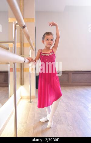 the charming little girl dreams of becoming a ballerina. The girl in the pink dress is dancing, holding on to the bar.Baby girl is studying ballet. Stock Photo