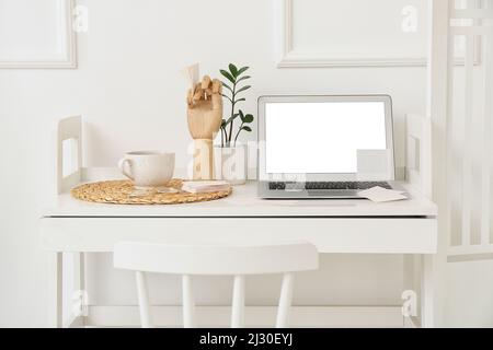 Laptop with sticky notes, wooden hand, houseplant and cup of tea on table near light wall Stock Photo