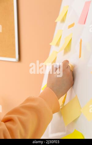 Woman writing on sticky note attached to flipchart near beige wall, closeup Stock Photo