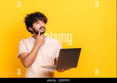 Handsome charismatic indian or arabian curly guy in basic t-shirt, holding open laptop in hand, looking away thoughtfully, planning, dreaming, standing over isolated orange background Stock Photo