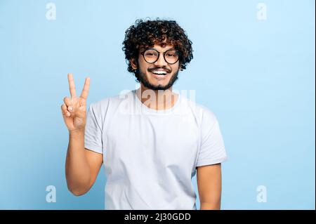 Charismatic handsome positive indian or arabic guy with glasses, freelancer or creative designer, stands on a blue isolated background, shows a PEACE sign, looks at the camera, smiles friendly Stock Photo