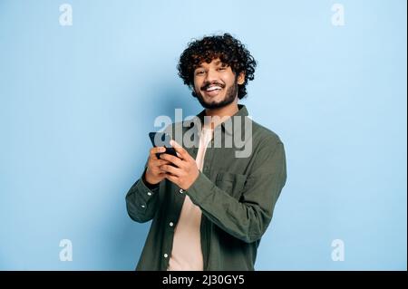 Happy handsome amazed stylish indian or arabian guy, using smartphone, chatting online, texting message, browsing internet, social media, looks at camera, stands on isolated blue background, smiling Stock Photo