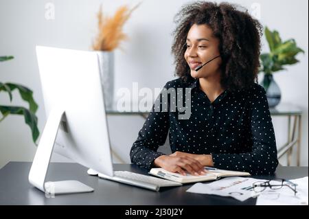 Positive beautiful confident african american woman with headphones, hotline operator, support worker, online consultant sits at computer in modern office, talking to a client on video call, smiling Stock Photo