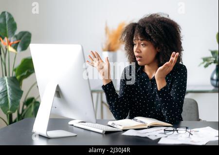 Surprised upset young adult curly haired african american business woman, manager or broker sits at a table, looks at computer, shocked by the news or messages, suffered financial losses, puzzled Stock Photo