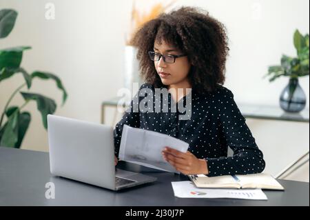 Focused smart purposeful young adult african american business woman, manager or office worker, in stylish clothes, sits at her desk, looks at laptop screen, works with documents and charts Stock Photo