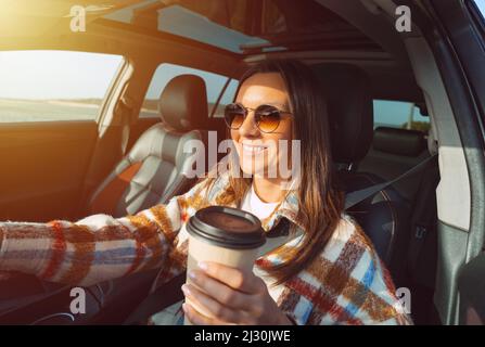 Happy smiling beautiful woman holding coffee cup and driving a car on a sunny day Stock Photo